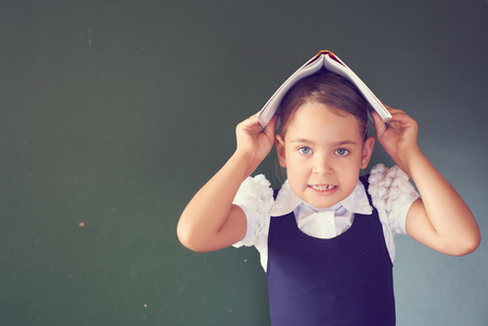 Pretty schoolgirl in a school dress stands holding an open book on her head near at the blackboard.の写真素材