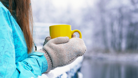North hipster girl relaxing on the pier at the frozen lake. Woman drinking hot tea from yellow cup and enjoing panorama lake view. Happiness, winter holidays, tourism, travel and people concept.の写真素材