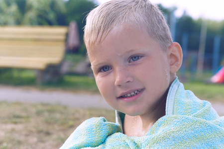 Little Caucasian boy basking after swim in the lake. The boy covered himself with the towel and sits on the shore of the lake .の写真素材