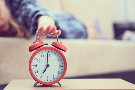 Young girl lies on the couch and stretches her hand to the red alarm clock to turn it off. Late wake up.の写真素材