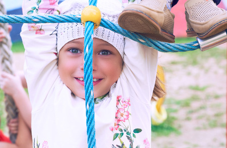 Portrait of cute Caucasian litte girl wearing white dress with flowers hanging on monkey bars on a...