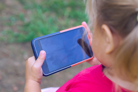 Baby girl holding a mobile phone. Little Caucasian girl playing with smartphone in summer park. Child learning how to use modern electronic devicesの写真素材