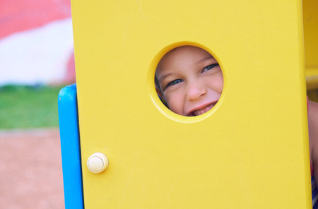 Face of small Caucasian boy looking through a hole in a play equipment outdoors. Orange background. Happy childhood conceptの写真素材