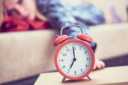 Young girl lies on the couch and stretches her hand to the red alarm clock to turn it off. Late wake up.の写真素材