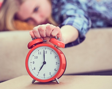 Young girl lies on the couch and stretches her hand to the red alarm clock to turn it off. Late wake up.の写真素材