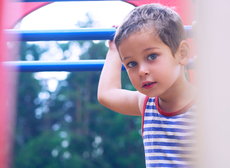 Little Caucasian boy in striped t-shirt hang the blue bar by his hand to exercise at out door...