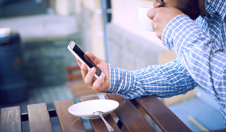 Modern hipster businessman in glasses drinking coffee in the city cafe during lunch time and using mobile phone.の写真素材
