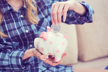 Young girl sitting on couch and putting litecoin in piggybank.の写真素材