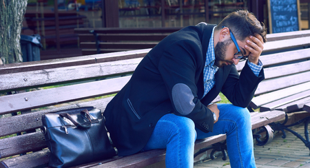 Young man with sad facial expression sitting on a bench in the park. Office worker lost his job. Middle aged man despair economic crisis.の写真素材