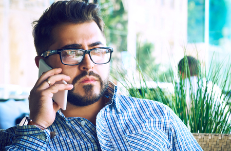 Concentrated businessman using mobile phone. Arabic businessman in glasses and beard having a phone talk seateing in the terrace of modern lounge cafe at his work break.の写真素材
