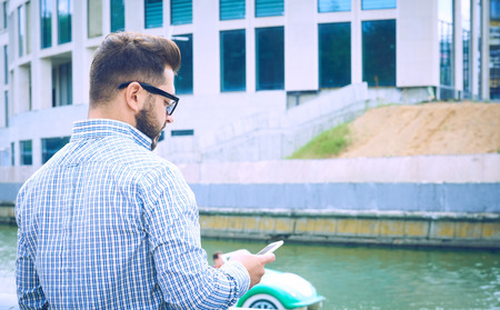 Back view. Hipster man is standing on the embankment of the river and using smartphone. Guy in denim...