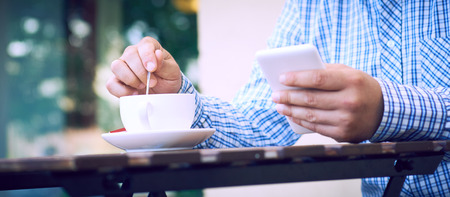 Always in touch with his colleagues. Close-up of man holding mobile phone and coffee cup while sitting on the terrace of the summer cafe.の写真素材