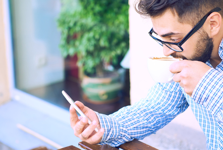 Modern hipster businessman in glasses drinking coffee in the city cafe during lunch time and using mobile phone.の写真素材