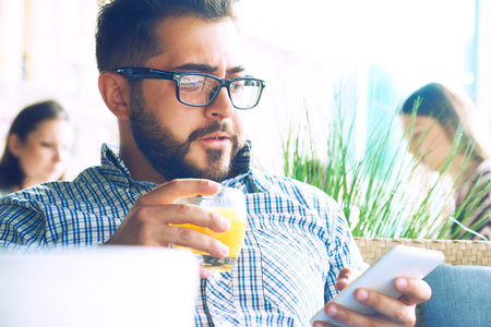 Handsome man with glasses drinking juice and looking at mobile phone in cafe.の写真素材