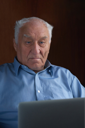Senior grey-haired man using a laptop sitting in chair at home.の写真素材