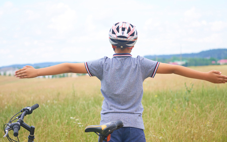 Back view of teenager boy in protective helmet spread his arms out like a bird standing next to his bike in park on summer day. Childrens outdoor activities in summer.の写真素材