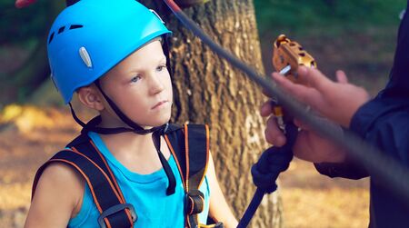 Cute little boy in blue shirt and helmet having fun at the adventure park, holding ropes and...