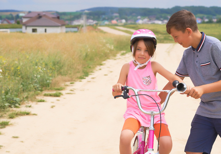 Youth brother teaching his younger sister to ride a bike. Little girl in a pink protective helmet on a pink bike rides with the support of her brotherの写真素材