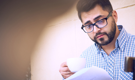 Young businessman in casual clothes and eyeglasses is studying document while drinking coffee on the terrace of a summer cafe.の写真素材
