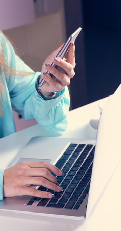 Young businesswoman having business call in office. Woman holds a smartphone in her hand and looks at the screen with her second hand typing on a laptop.の写真素材