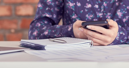 Close-up of female hands using smart phone while working on computer at modern office interior, businesswoman typing text message on her cellphone at loft interior.の写真素材