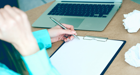 Stressed business woman write in clipboard in the surrounding of laptop and crumpling paper at her table in the office. Woman trying to come up with the right solutionの写真素材