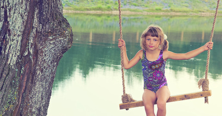 Beautiful little Caucasian girl in a blue swimsuit sitting on the swing in the forest by the lake. Color image of vintage styleの写真素材