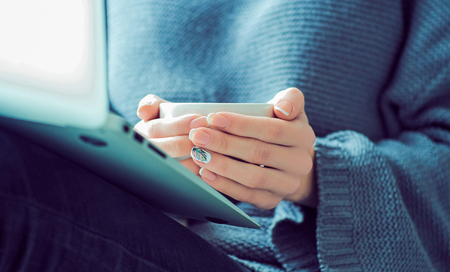 Girl sending a message, e-mail, woman typing something on the laptop in her lap sitting near window and holding cup of coffee close-up.の写真素材