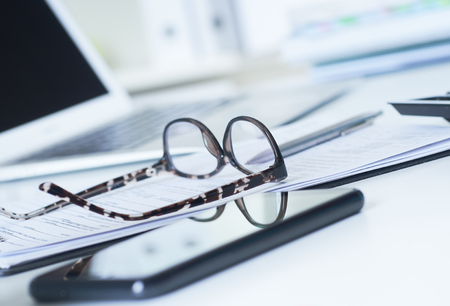 Closeup of white desktop with laptop, spectacles, notepads, pen, smartphone, clipboard.の写真素材