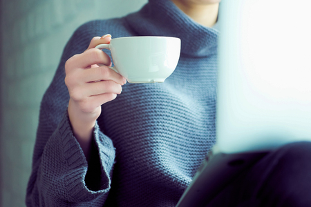 Girl sending a message, e-mail, woman typing something on the laptop in her lap sitting near window and holding cup of coffee close-up.の写真素材