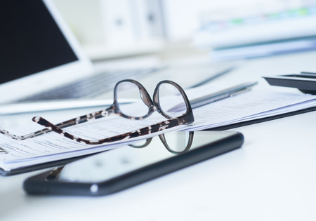 Closeup of white desktop with laptop, spectacles, notepads, pen, smartphone, clipboard.の写真素材