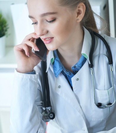 Young female therapeutist doctor sitting in front of working table with laptop and documents on it and calling on the phone.の写真素材