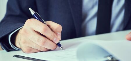 Hand of businessman in suit filling and signing with blue pen partnership agreement form clipped to...