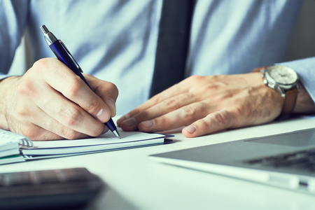 Businessman hand writing note on a notebook. Business man working at office desk. Close up of empty notebook on a blackboard with office supplies.の写真素材