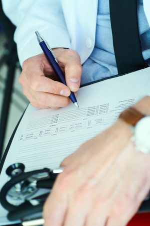 Male doctor on duty in white coat with pen in hand filling prescription or checklist document close up. Selective focus on pen, health and medical concept.の写真素材