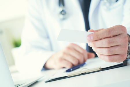 Male physician hand holding and giving white blank calling card closeup in office. Contact information exchange, introducing gesture at formal meeting, personal or family doctor conceptの写真素材