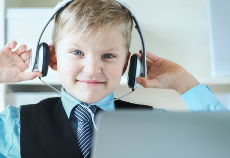 Cute 6 year old boy in suit listening to music or audio tutorial on headphones at the office background.の写真素材