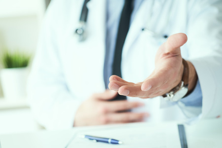 Male doctor making welcome gesture, politely inviting patient to sit down in medical office. Photo with depth of field.の写真素材