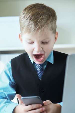 Smart little boy in suit working on laptop and touching the phone screen emotionally.の写真素材