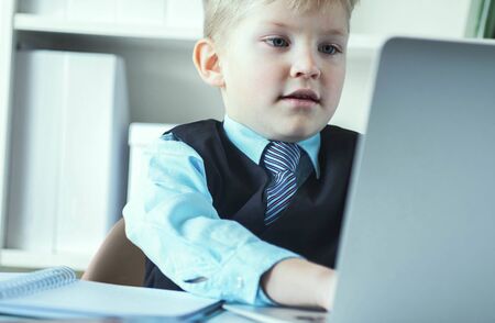 Small cute boy in business suit with a tie works at computer at office. Small business man, little...