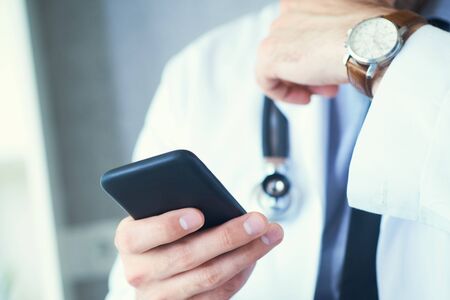 Male doctor in white coat is using a modern smartphone device with touch screen. Doctor hands with mobile phone.の写真素材