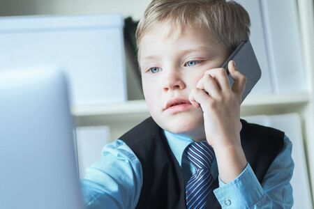 Little confident executive businessman boss boy in office talks on the phone sitting at the desk with laptop.の写真素材