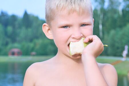 Little Caucasian blonde boy eating an pear on a hot summer day at the beach.の写真素材