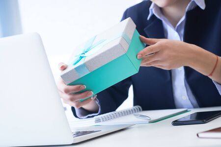 Christmas, any holiday. Office employee received a gift from her colleagues. Closeup woman hands holds gift box on office background.の写真素材