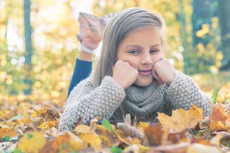 A portrait of a beautiful little girl lying on the ground with golden autumn leaves. Happy childhood, lifestyle, autumn fashion, beauty.の写真素材