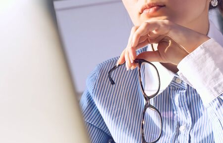 Thoughtful female office manager with spectacles in hand near the chin looking at laptop screen...