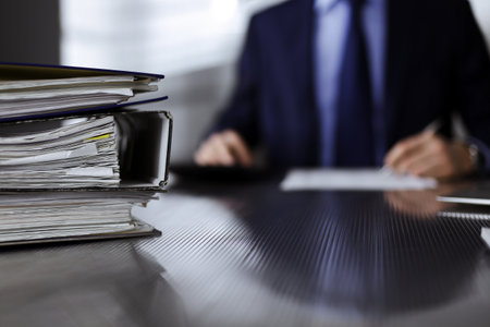 Binders with papers are waiting to be processed by man accountant in blue blazer staying at home during covid pandemic. Taxes and audit concept. Internal Revenue Service inspector checking financial document.の写真素材