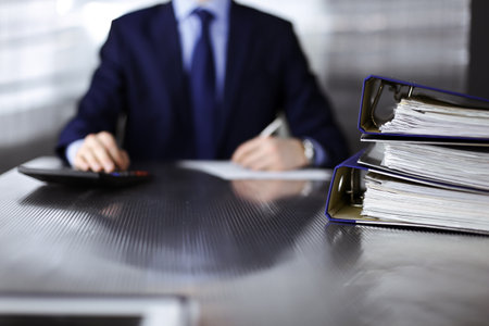 Binders with papers are waiting to be processed by man accountant in blue blazer staying at home during covid pandemic. Taxes and audit concept. Internal Revenue Service inspector checking financial document.の写真素材