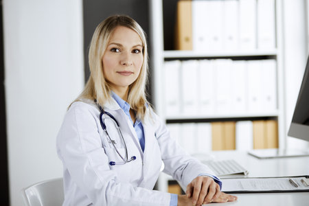 Friendly female doctor sitting in clinic at her working place. Portrait of cheerful smiling physician. Perfect medical service in hospital. Medicine and healthcare conceptの写真素材