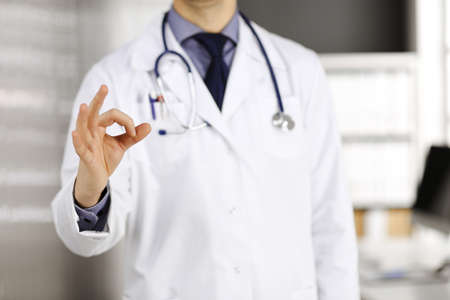 Unknown male doctor standing straight with OK sign in clinic near his working place, closeup. Perfect medical service in hospital. Medicine and healthcare conceptの写真素材